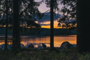 Skymningsljus över en sjö och skog med ett tält vid vattenkanten. Tre personer sitter på huk vid en eld.