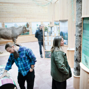 En kvinna i grön jacka och en man med skägg och blårutig skjorta läser och tittar på utställningen Skogen bortom träden.