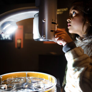 En flicka med bruna ögon och mörkbrunt hår tittar i ett mikroskop.